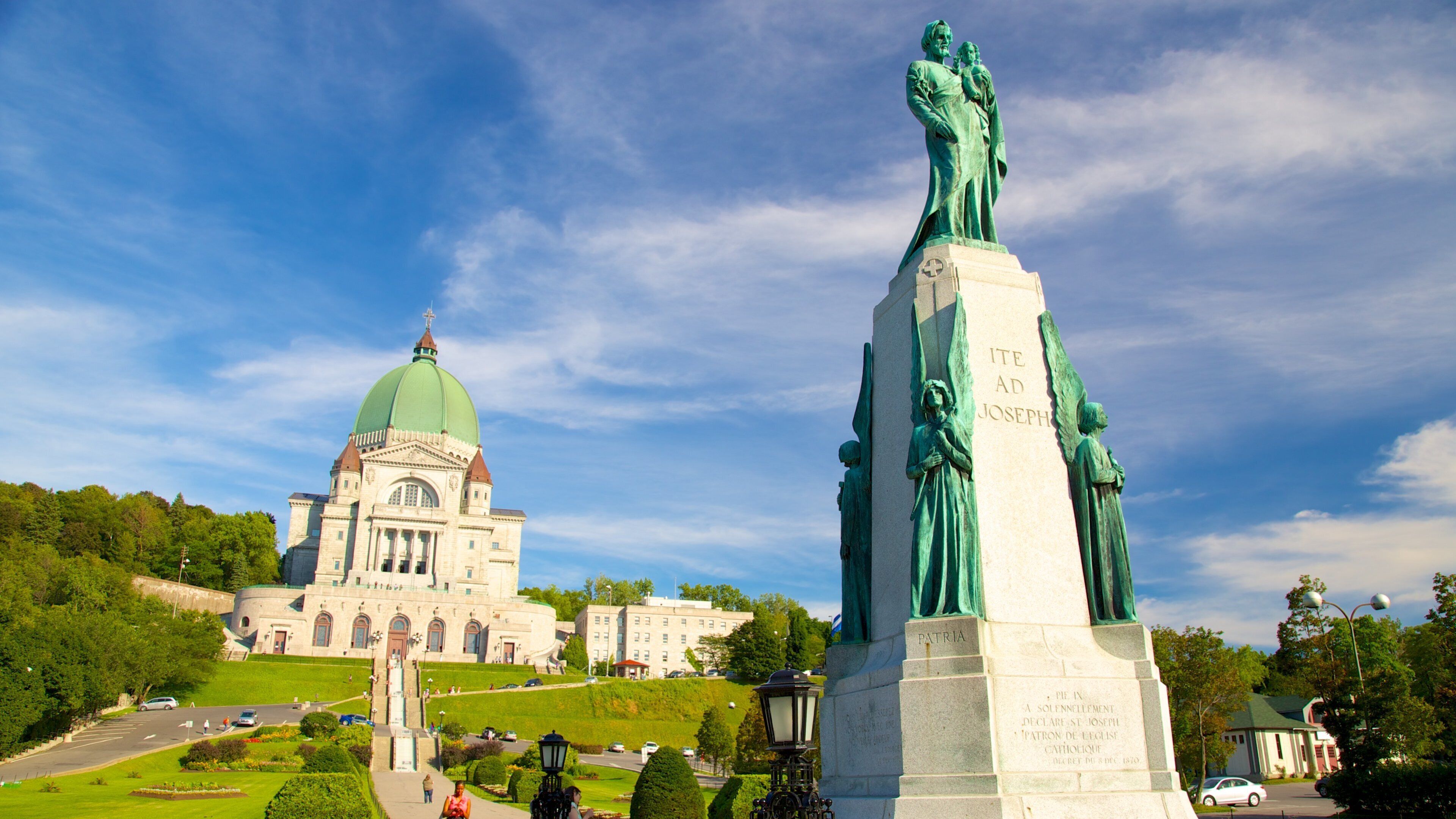 A statue of a religious figure with inscriptions, a large green-domed building, and a well-maintained park with pathways.