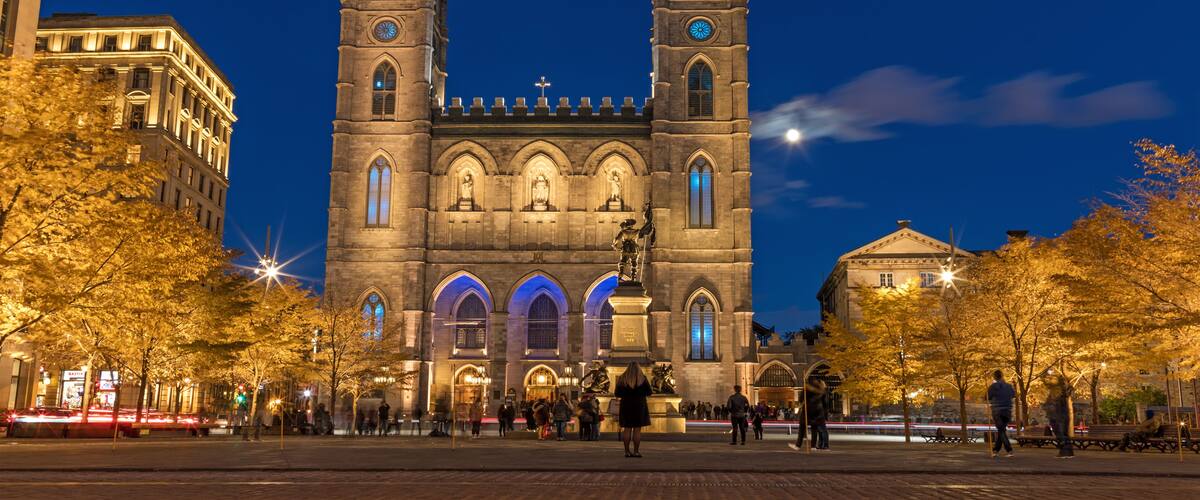 Una catedral histórica con dos torres gemelas y un gran arco central, rodeada de árboles otoñales y una calle concurrida.