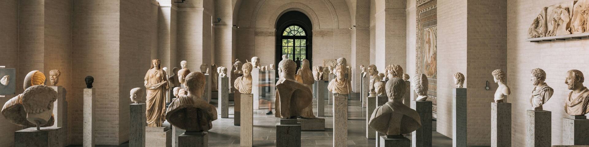A museum with classical sculptures on pedestals, arched ceilings, and a large window.