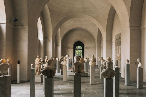 Ein Museum mit klassischen Skulpturen auf Podesten, gewölbten Decken und einem großen Fenster.