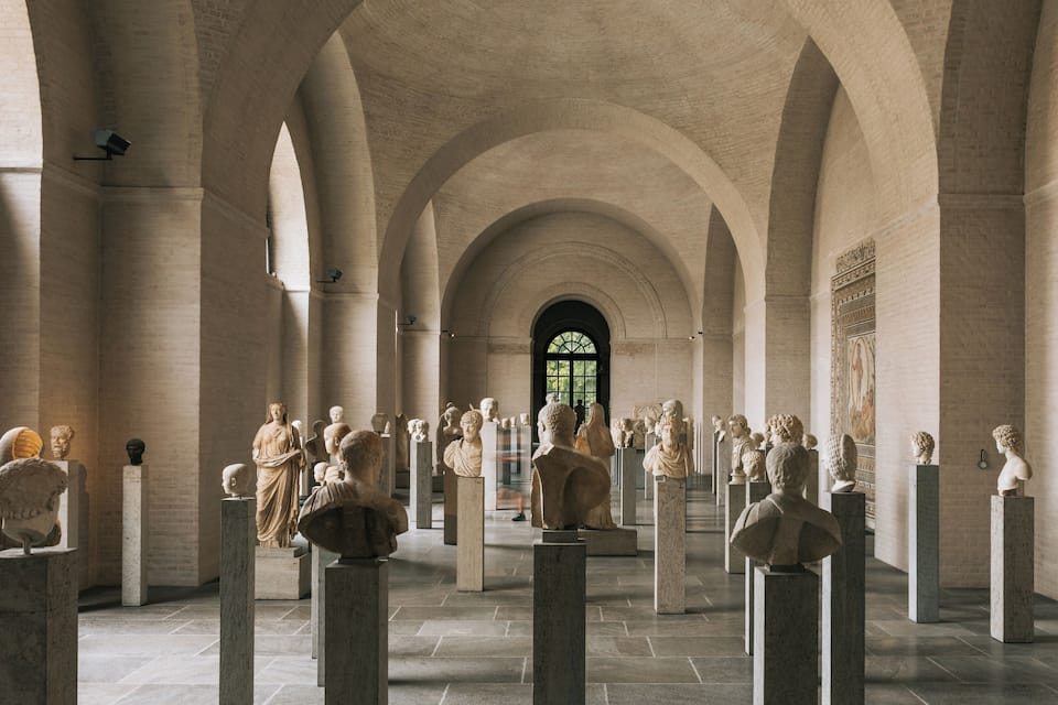 A museum with classical sculptures on pedestals, arched ceilings, and a large window.