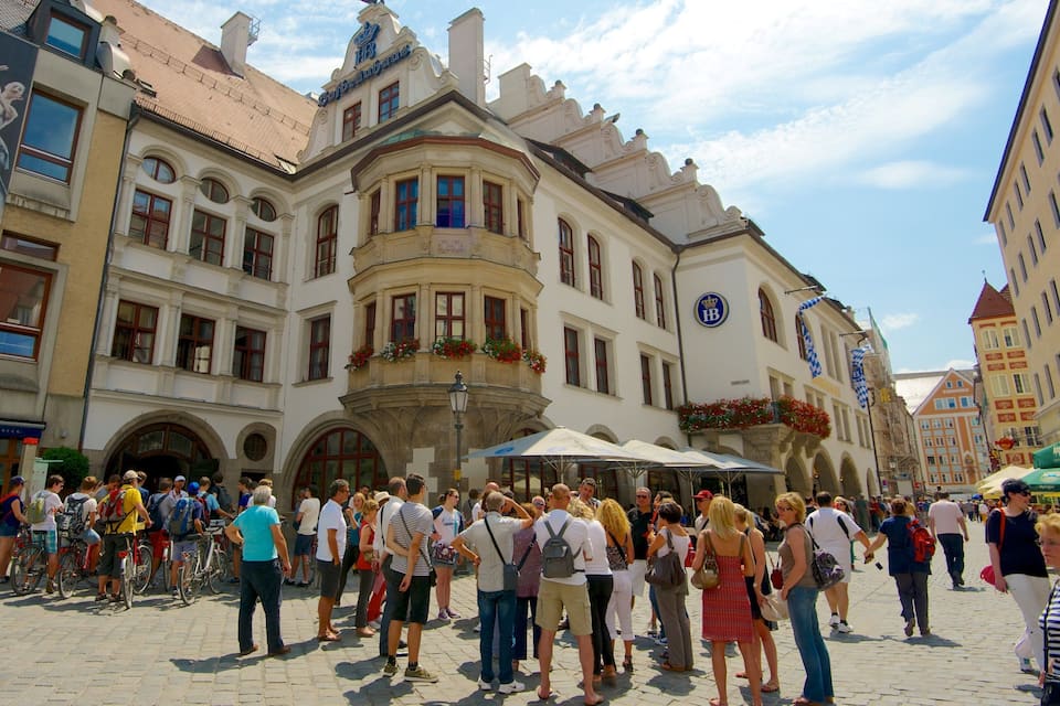A crowded square with historic buildings, a prominent 'HB' sign, and people gathered around.