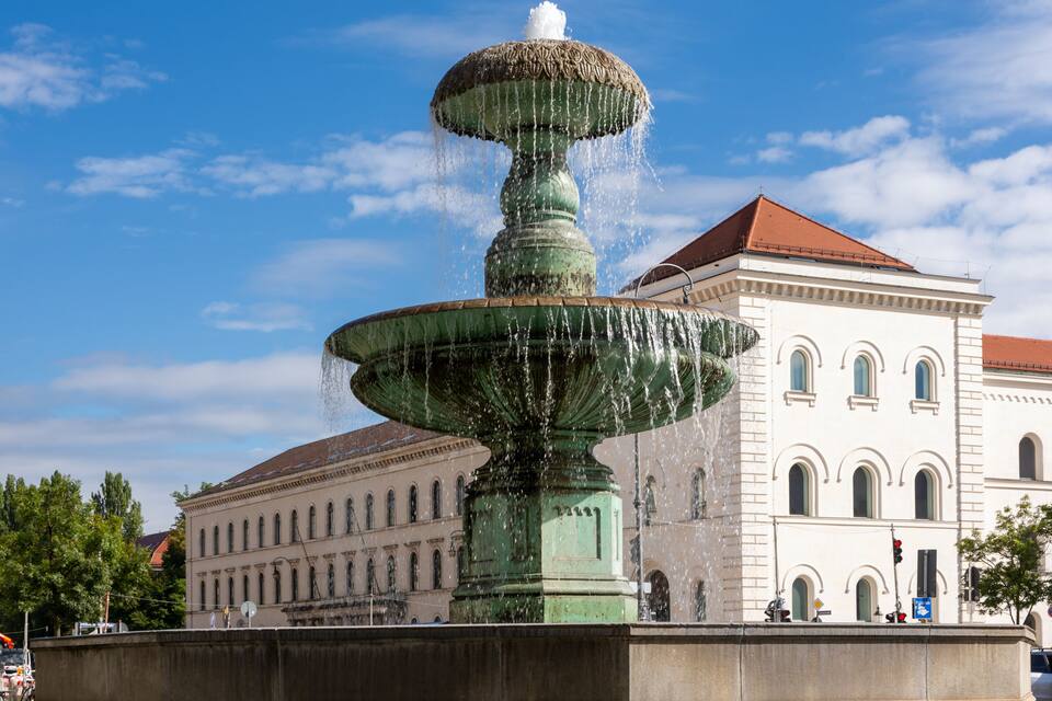 A multi-tiered fountain with water jets in front of a classical building with arched windows.