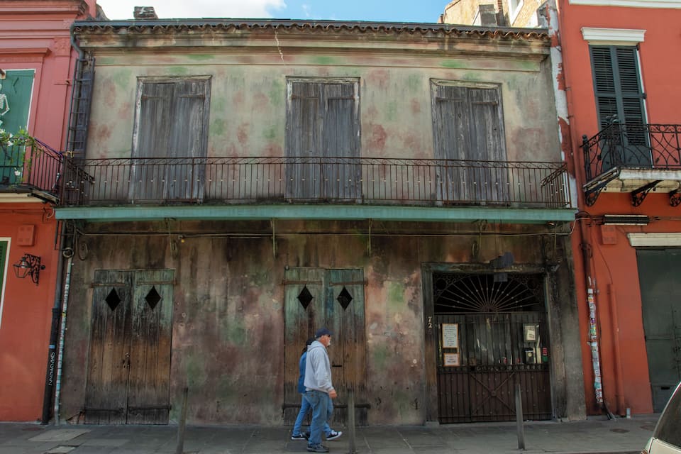Um homem passa por um edifício desgastado, com persianas verdes e uma varanda.