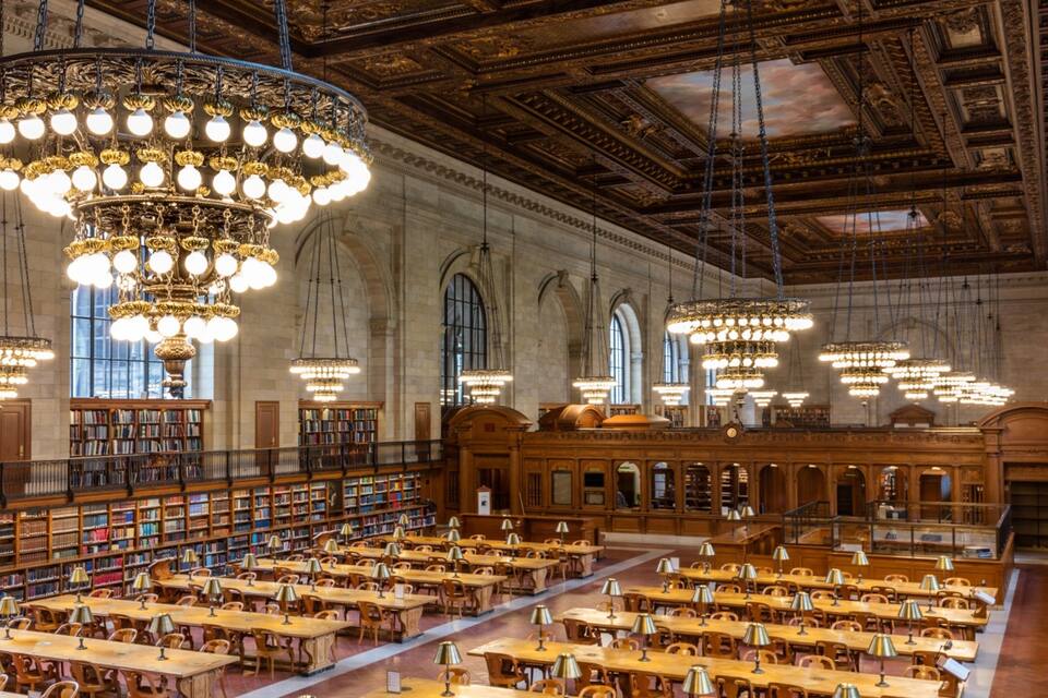 Una biblioteca imponent amb lluminàries ornamentals, escriptoris de fusta i prestatgeries plenes de llibres.