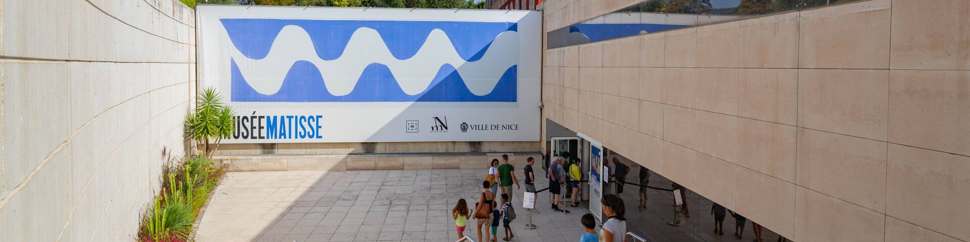 A modern building with a large blue and white mural, a red building in the background, and people walking on the stairs.