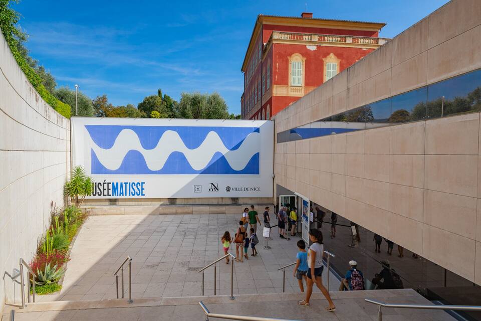 Ein modernes Gebäude mit einer großen, blau-weißen Wandmalerei, ein rotes Gebäude im Hintergrund und Personen, die die Treppe hinaufgehen.
