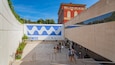 A modern building with a large blue and white mural, a red building in the background, and people walking on the stairs.
