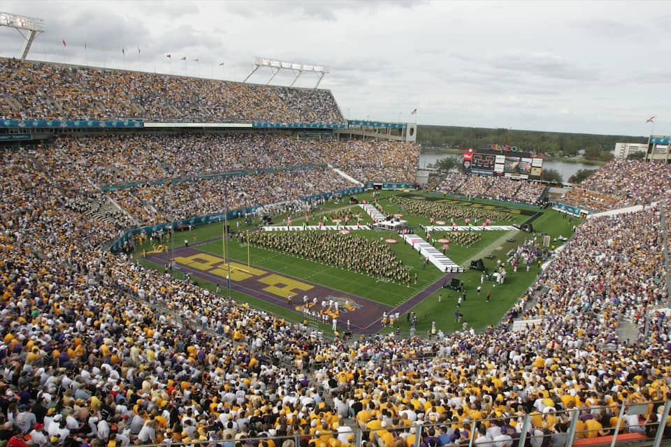 A football stadium filled with spectators, a marching band, and a large crowd of people wearing yellow and white uniforms.