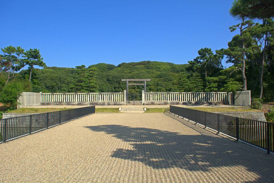 A paved pathway leading to a traditional Japanese torii gate surrounded by lush greenery and a clear blue sky.
