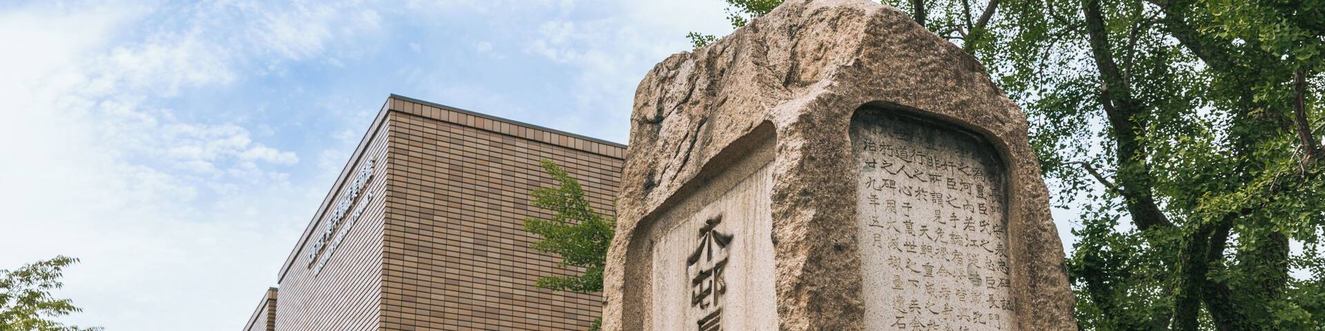 A stone monument with inscriptions, a modern building, and trees in the background.