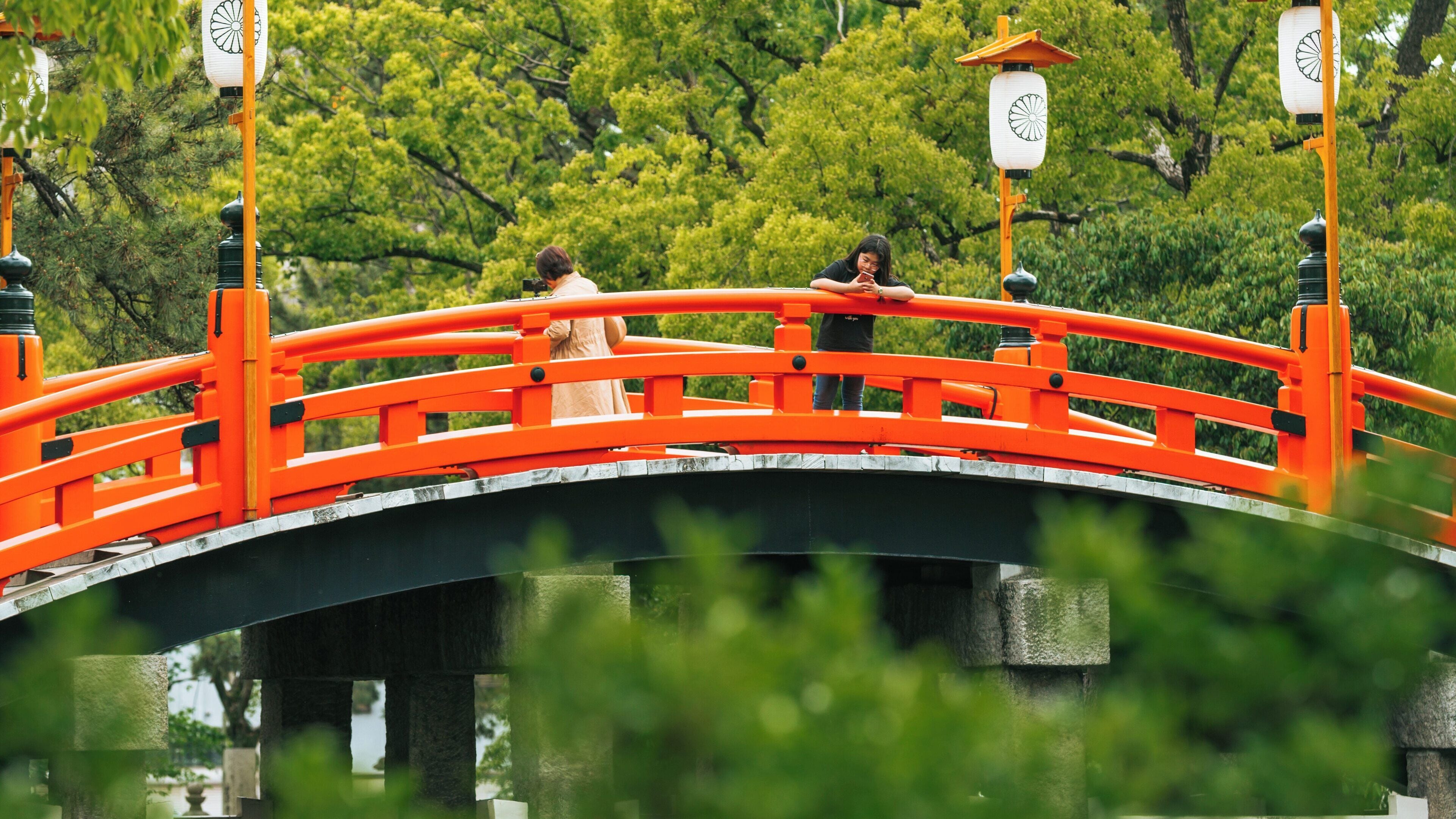 Visitors explore the iconic red bridge at Sumiyoshi Taisha Shrine in Osaka, Japan during a sunny day, surrounded by lush green trees and traditional architecture