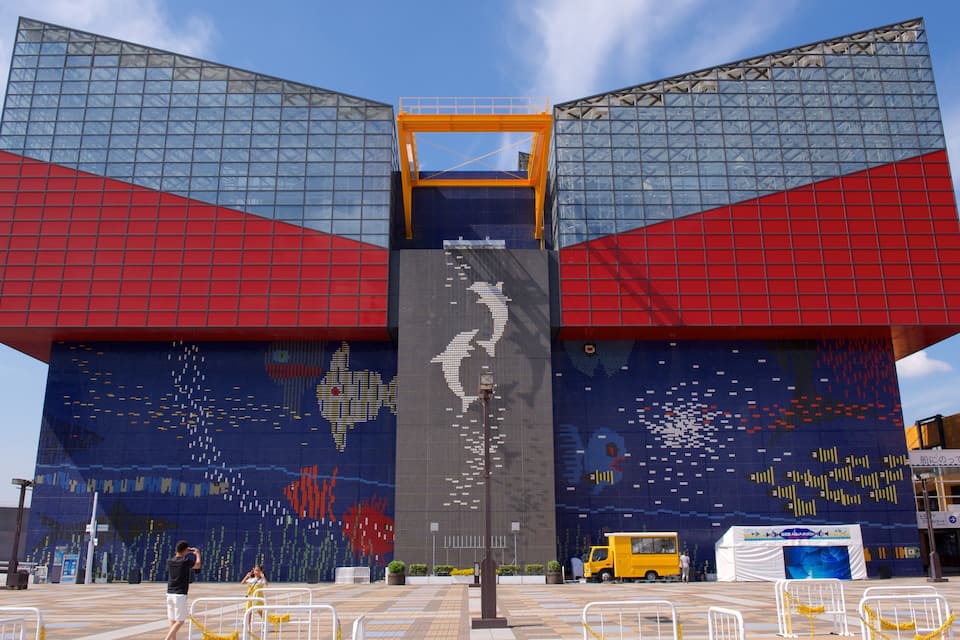 Un edificio grande con un murale a mosaico dai colori vivaci, una struttura centrale gialla e un cielo azzurro.