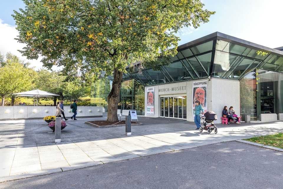 A modern building with a glass entrance, surrounded by trees and a paved area with benches.