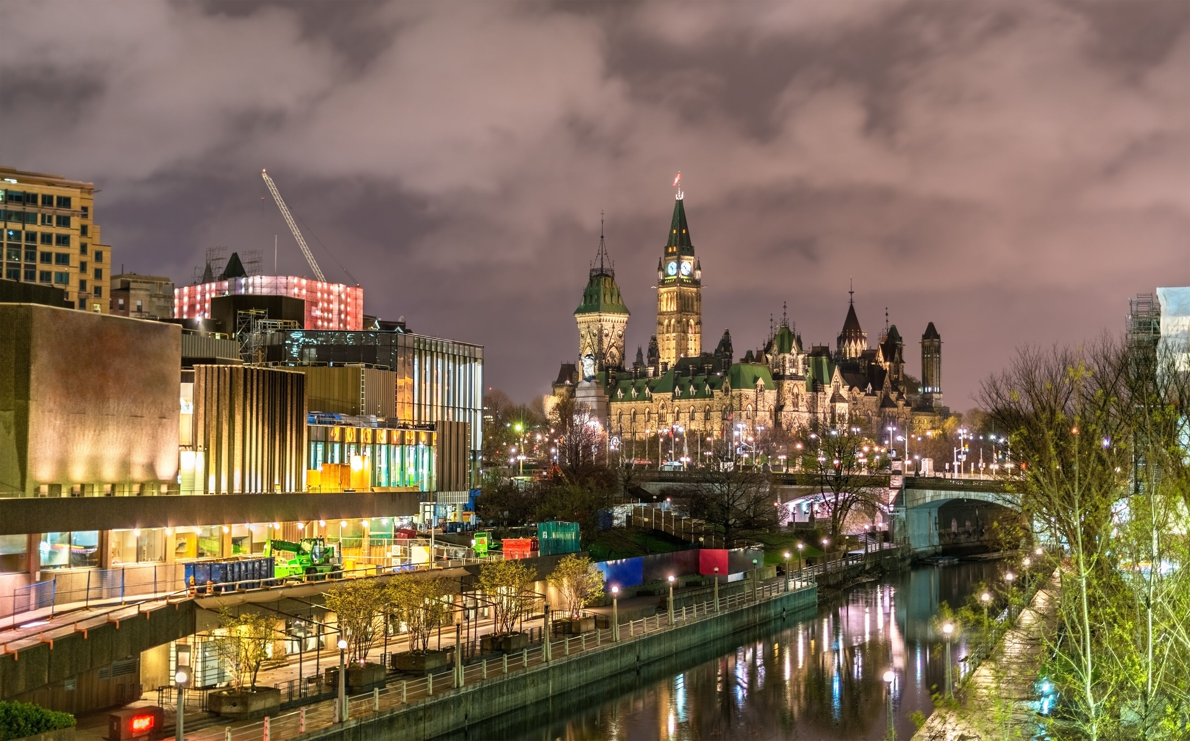 Un paisaje urbano nocturno con un edificio histórico destacado que presenta una torre con reloj, un río que refleja las luces y edificios modernos con fachadas iluminadas.