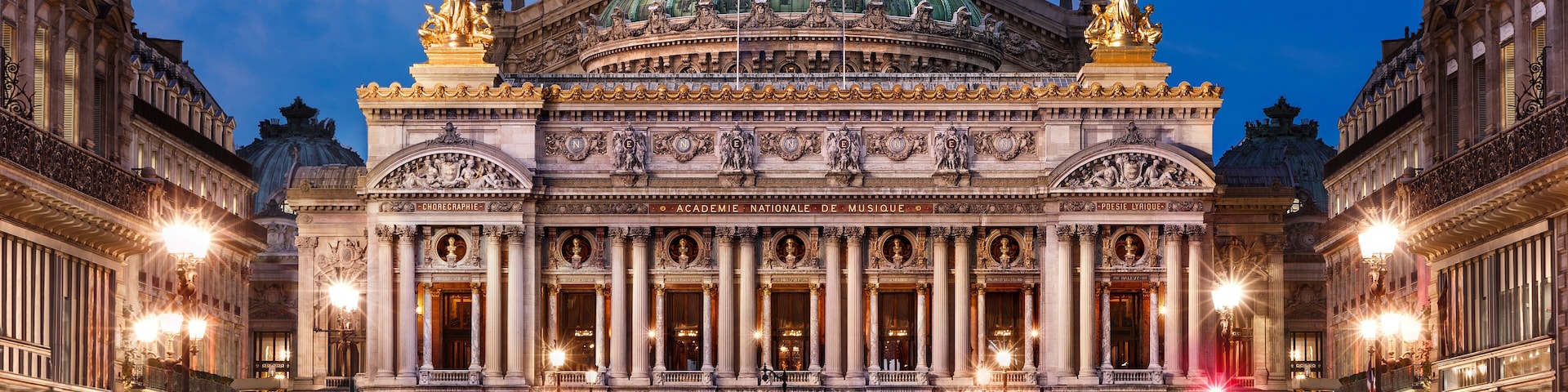 Um imponente teatro de ópera com uma grande cúpula e estátuas no telhado, rodeado por edifícios históricos.