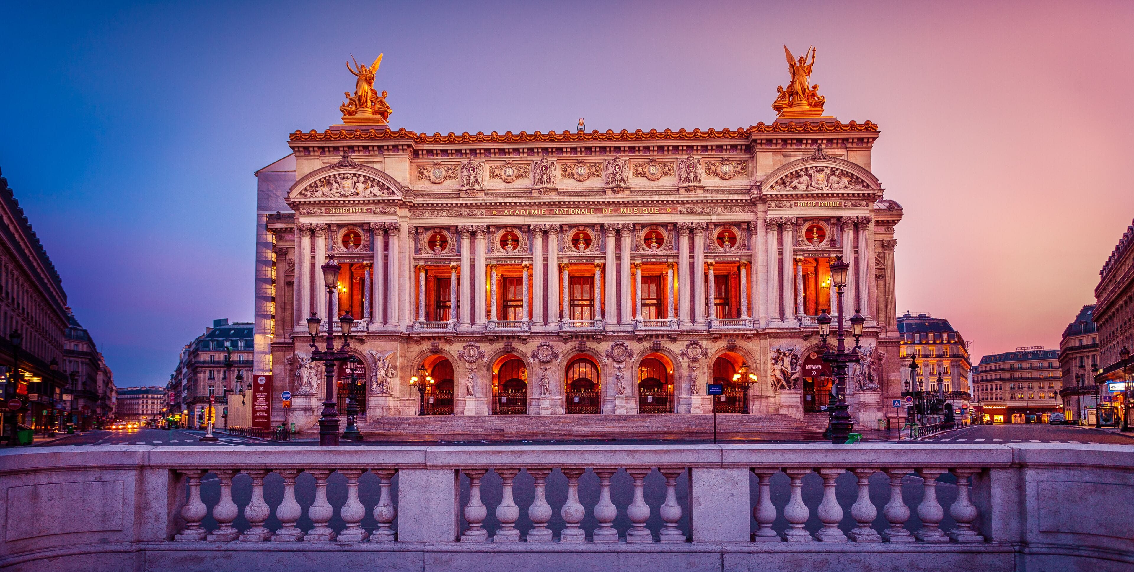 A grand, illuminated building with classical architecture, featuring statues and ornate details.