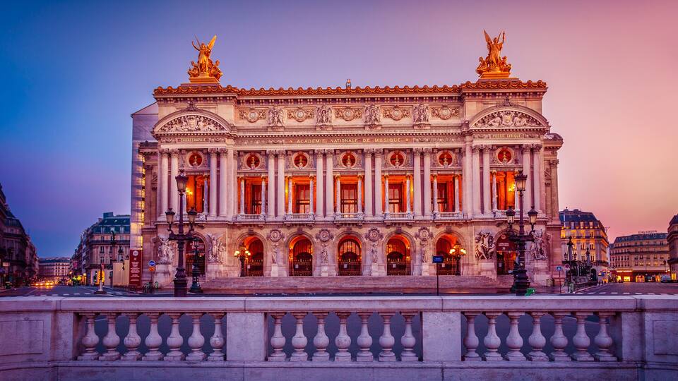 A grand, illuminated building with classical architecture, featuring statues and ornate details.