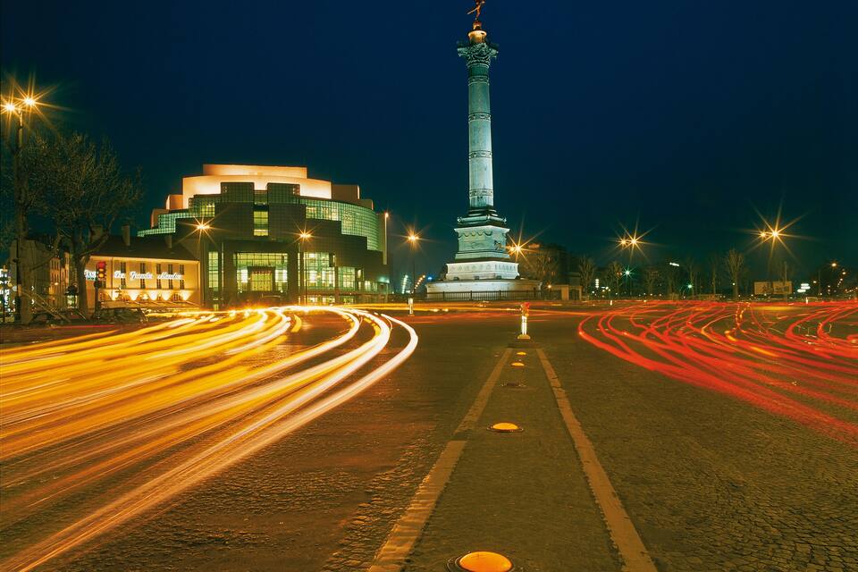 A nighttime cityscape with a prominent monument in the center, illuminated buildings, and light trails from moving vehicles.