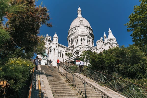 Um edifício imponente com cúpula e arquitetura detalhada, rodeado por vegetação exuberante e uma escadaria que leva até ele.