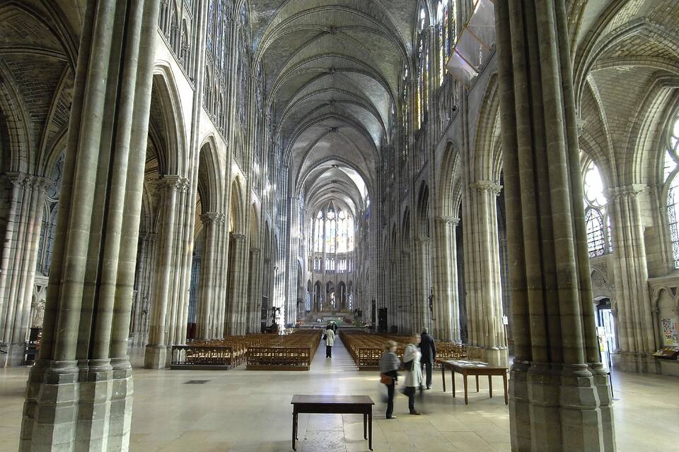 A cathedral interior with high vaulted ceilings, large stained glass windows, and rows of wooden pews.