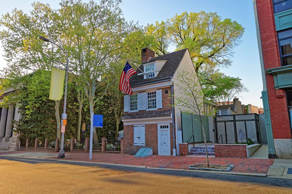 A historic brick house with a flag, surrounded by trees and a brick sidewalk.