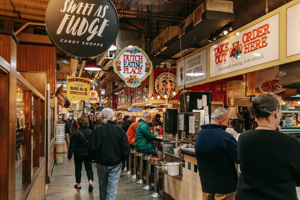 Un mercado techado concurrido con varios puestos de comida y letreros que anuncian artículos como 'Dutch Eating Place' y 'Take Out Here'.