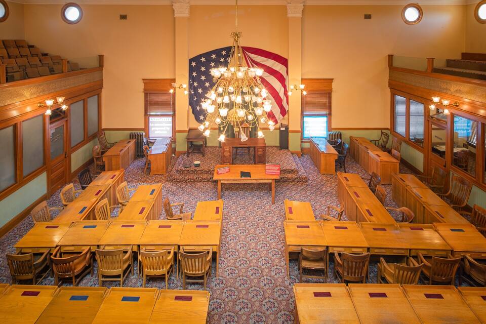 A grand hall with wooden tables and chairs, a large American flag, and a chandelier.