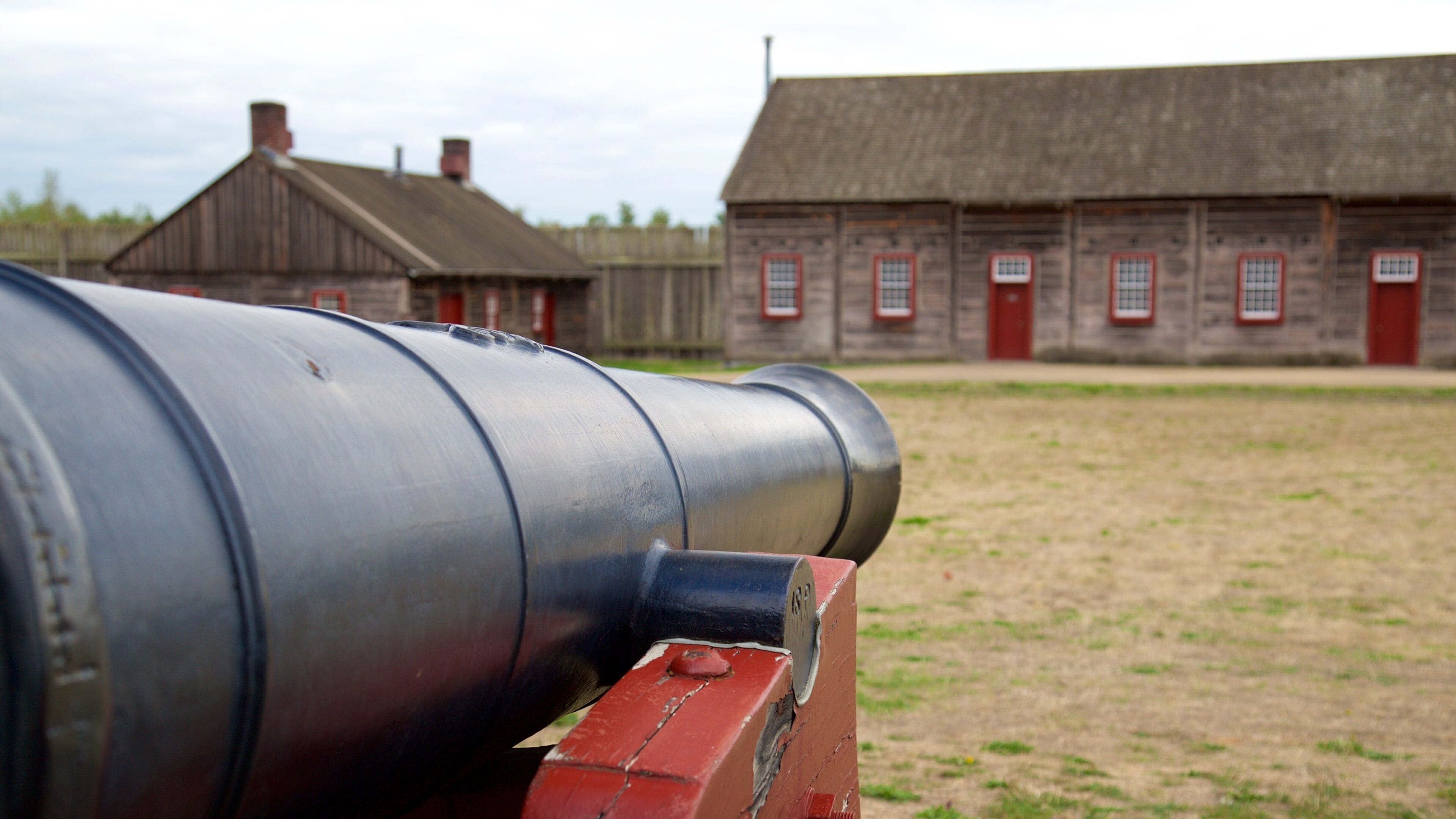 A large black cannon in the foreground with a red base, set against a backdrop of a wooden building with red doors and windows.