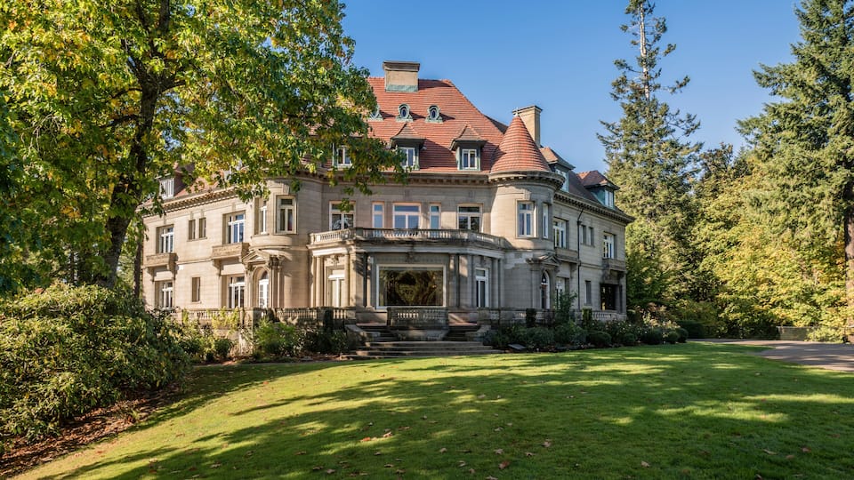 A large, historic building with a red-tiled roof, surrounded by lush greenery and trees.