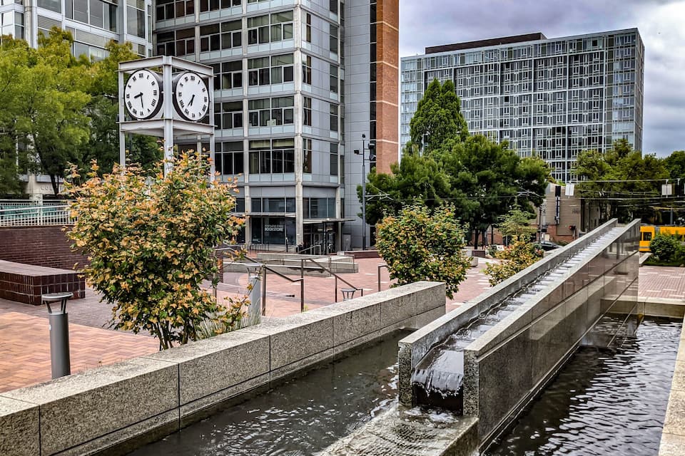 Un quartier urbain moderne doté d’un élément aquatique, d’une tour horloge et de bâtiments contemporains.