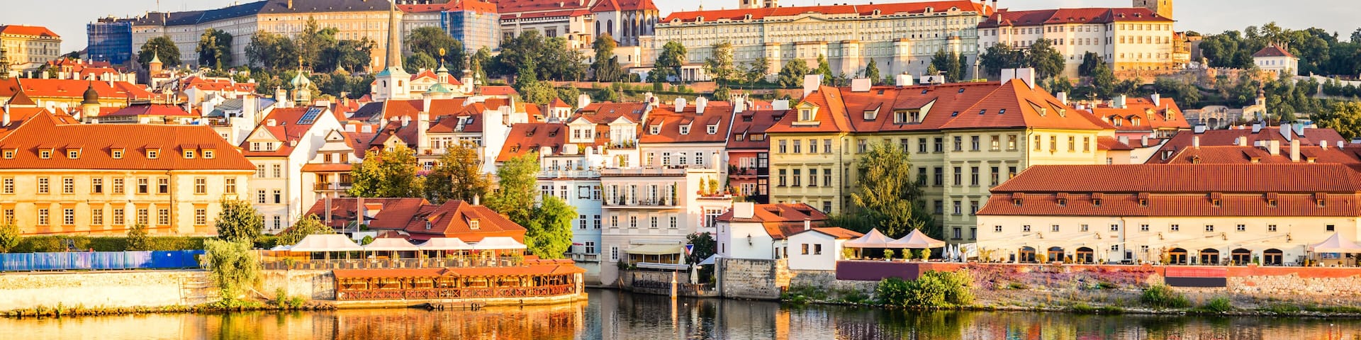 A historic cityscape with a prominent castle, a river reflecting buildings, and a clear sky.