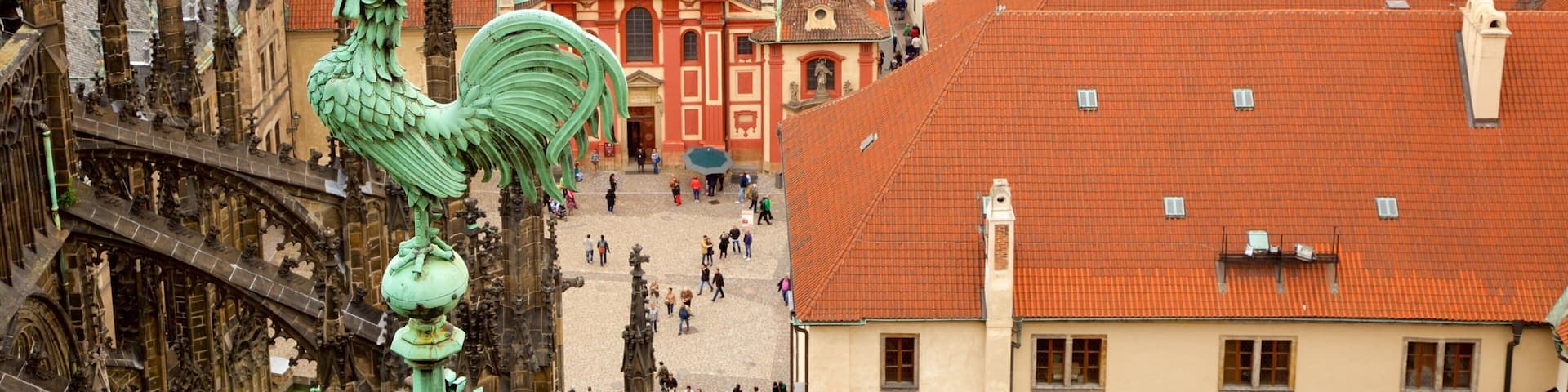 A historic town with red-tiled roofs, a prominent rooster statue, and a central square with people.