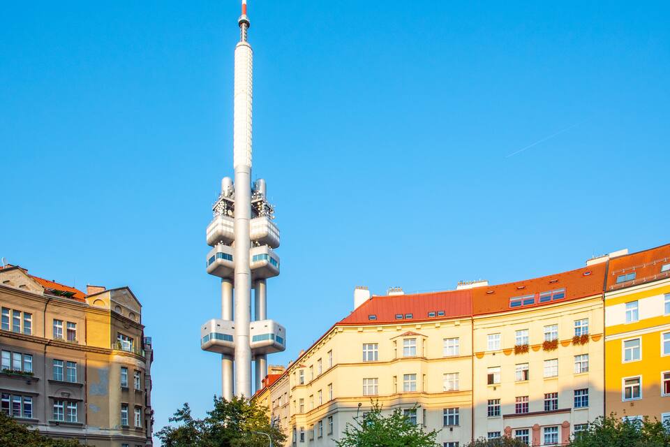 Uma torre alta e esguia com topo listrado em vermelho e branco, cercada por edifícios de vários andares com fachadas vermelhas e amarelas.