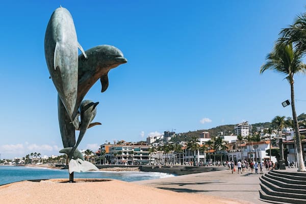 En stor delfin-skulptur på en strand med blå himmel, palmer og en by i baggrunden.