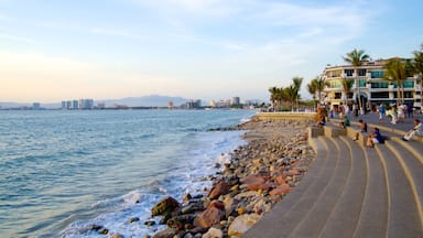 Un paseo marítimo costero con un litoral rocoso, mar en calma y un paisaje urbano al fondo.