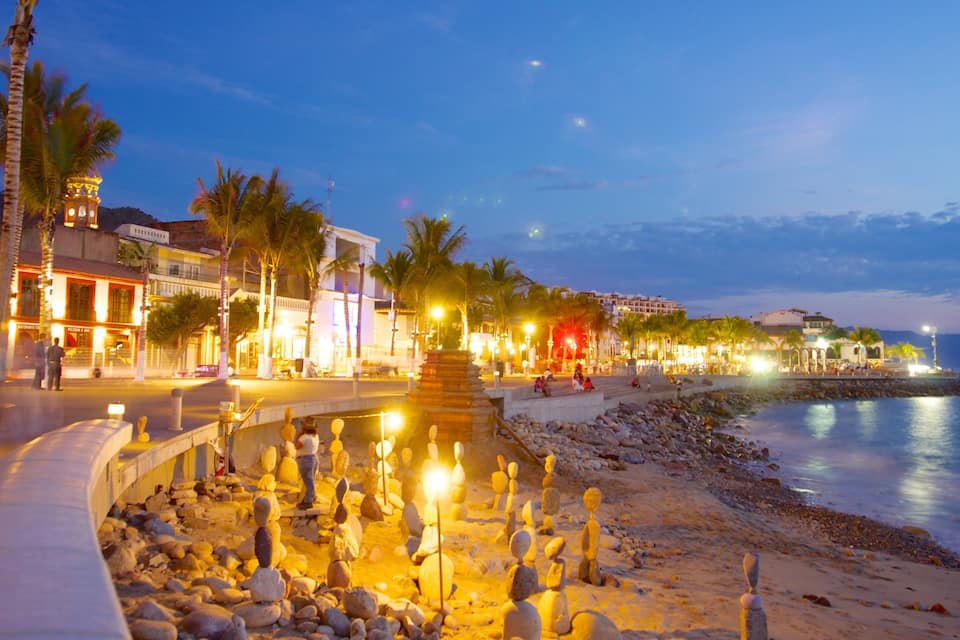 A beach at dusk with a rocky shoreline, palm trees, and illuminated buildings.