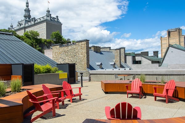 Un espace extérieur aménagé avec des chaises Adirondack rouges et des tables en bois, offrant une vue sur un bâtiment historique doté d’un clocher.