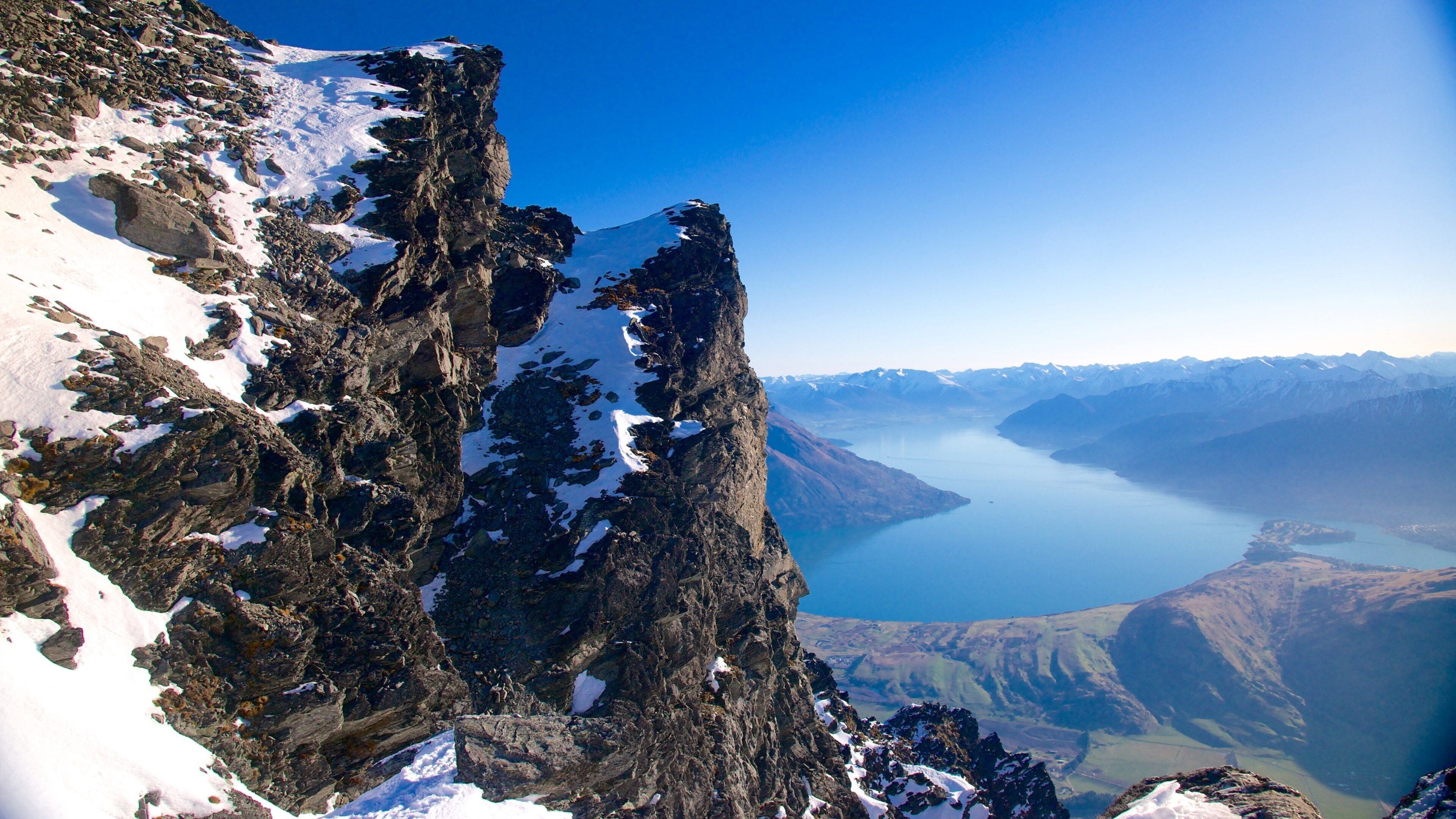 A rugged mountain peak with snow patches, a clear blue sky, and a distant lake.