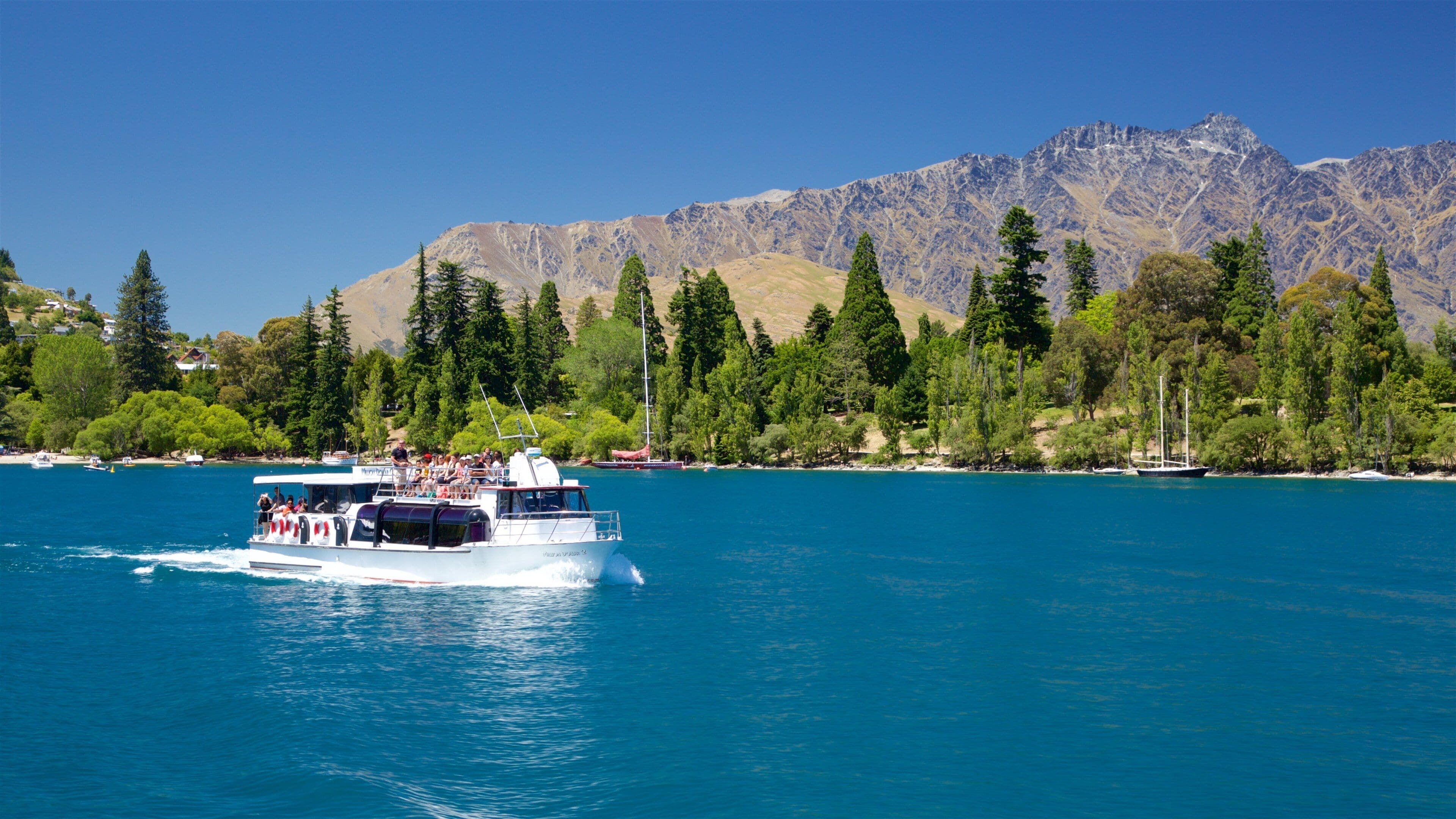 A boat with passengers on a clear blue lake, surrounded by mountains and dense green trees.