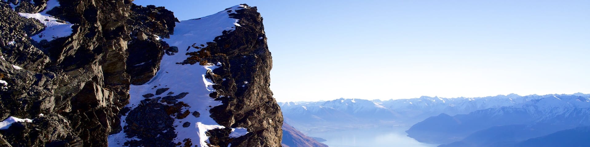 A rugged mountain cliff with snow patches, a deep valley, and distant snow-capped mountains.
