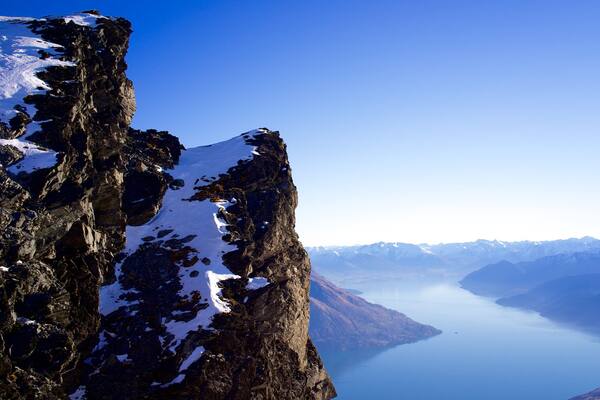 A rugged mountain cliff with snow patches, a deep valley, and distant snow-capped mountains.