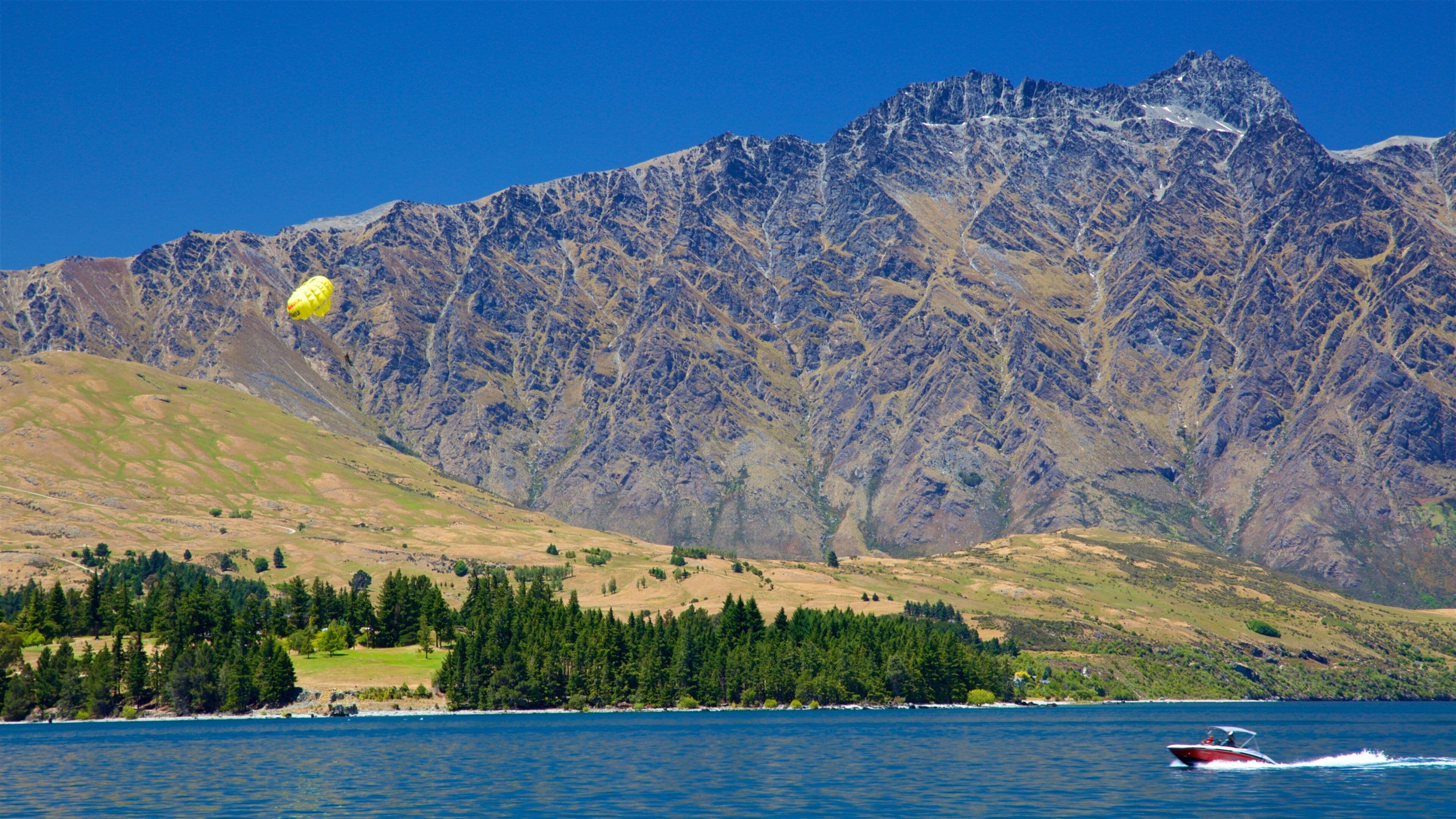 A lake with a mountain range in the background, a boat on the water, and a yellow parachute.