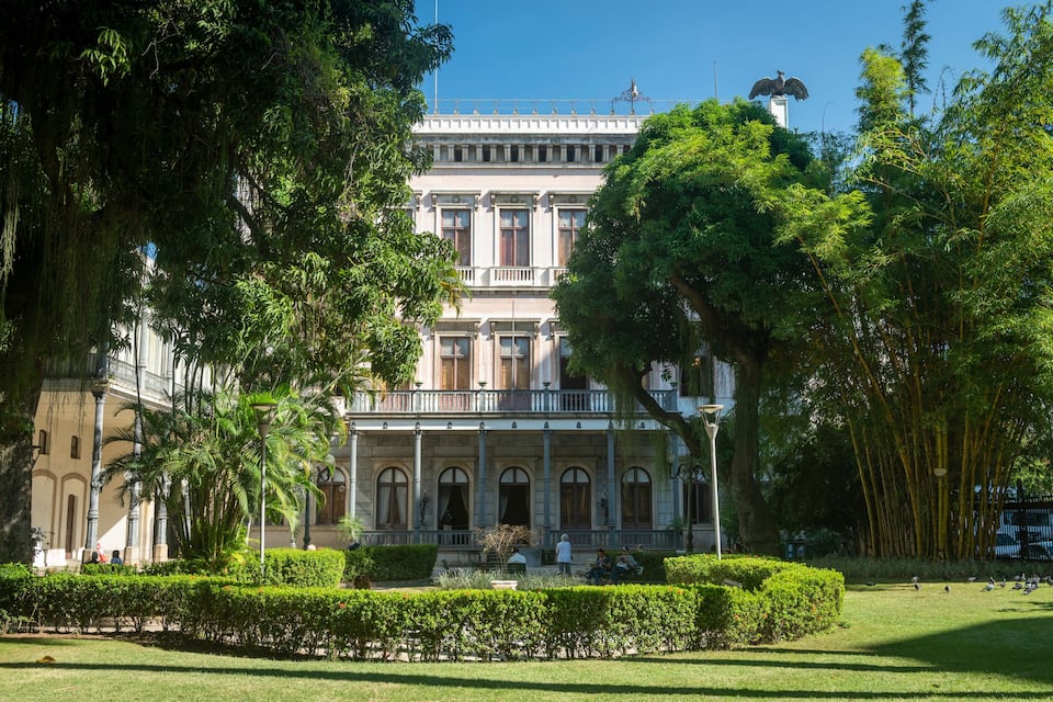 Un bâtiment historique doté d’un balcon, entouré d’une végétation luxuriante et d’arbres.