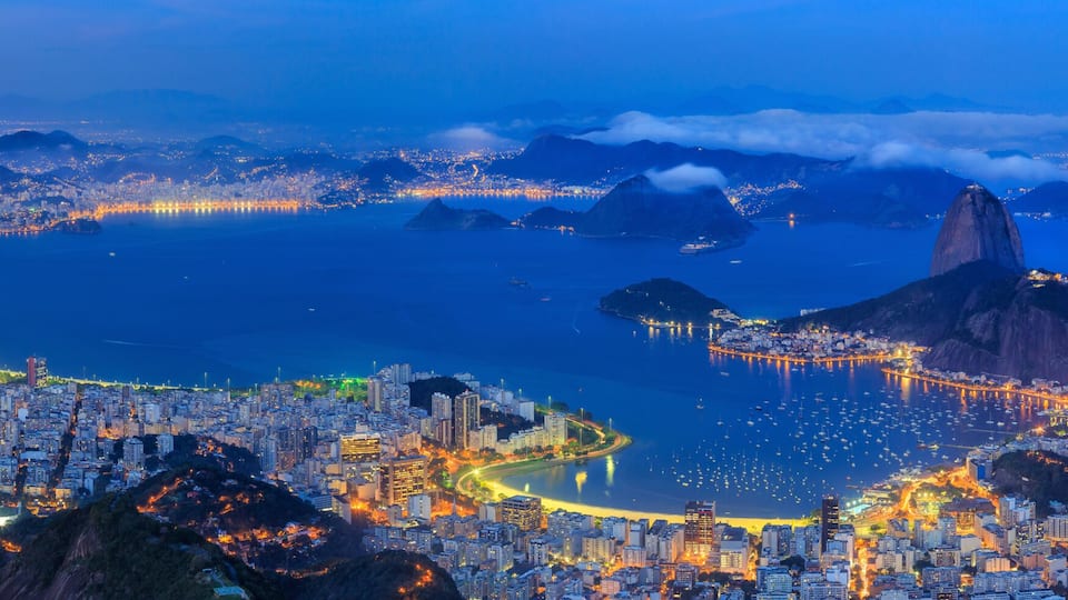 Vista panorâmica da cidade com uma cadeia montanhosa imponente, uma baía com muitos barcos e uma área urbana densa com edifícios iluminados.