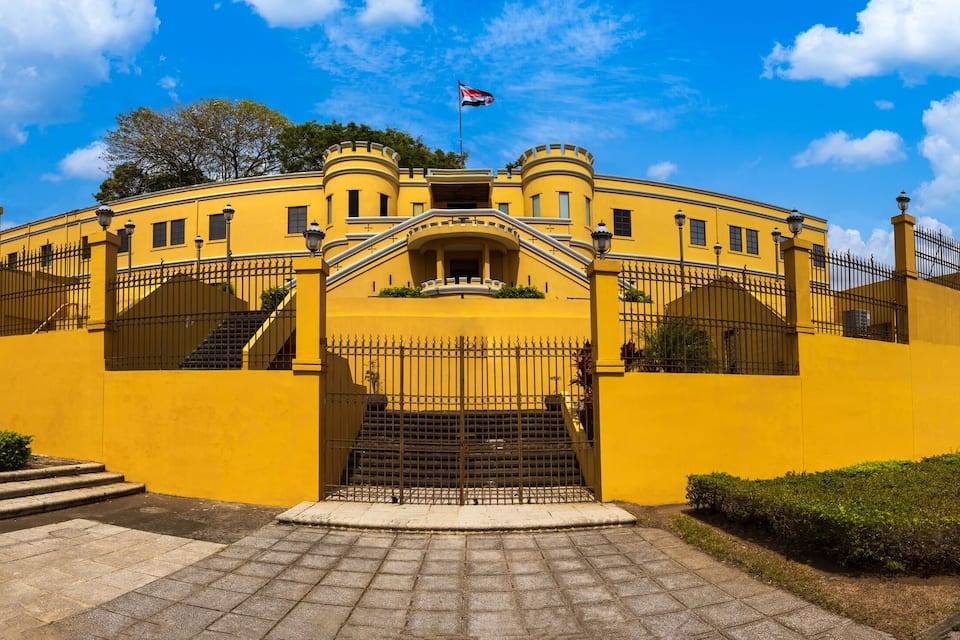A yellow building with a flag, steps, and a fence.