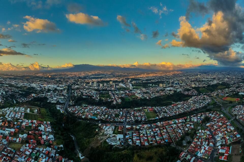 A panoramic view of a city with a prominent stadium, dense residential areas, and distant mountains under a partly cloudy sky.