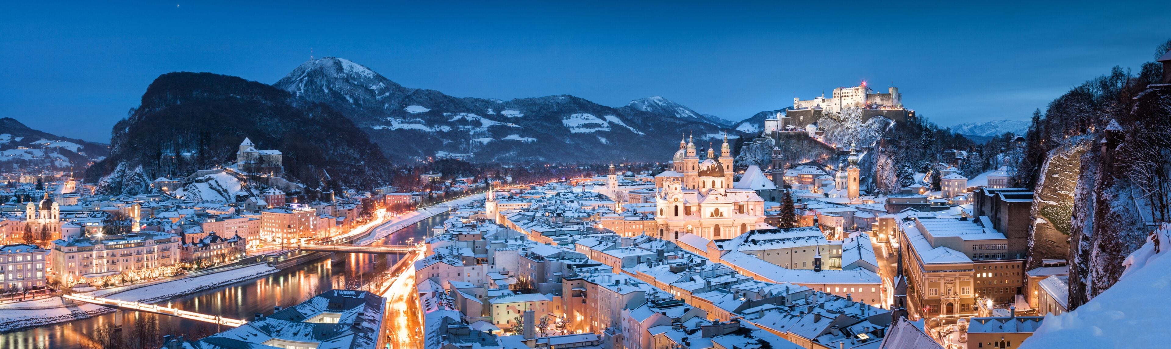 A snow-covered city with a prominent castle on a hill, a river running through the town, and mountains in the background.