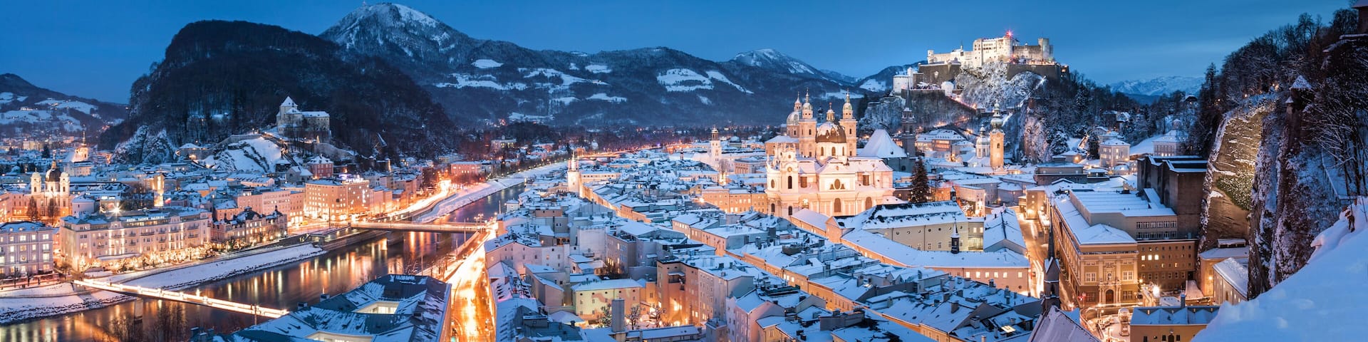 A snow-covered city with a prominent castle on a hill, a river running through the town, and mountains in the background.