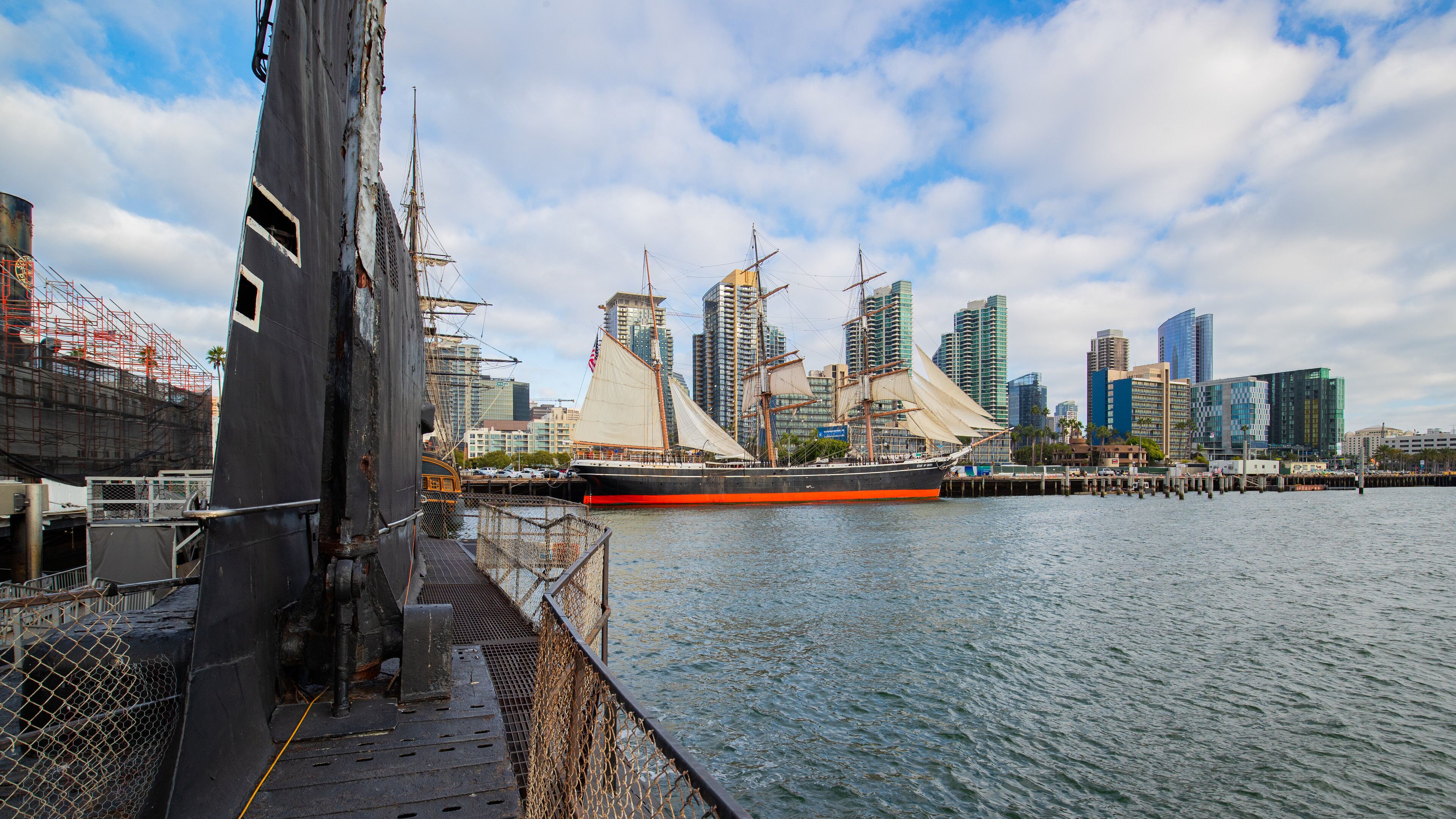 Um navio a vela histórico atracado em uma orla com um horizonte urbano moderno ao fundo.